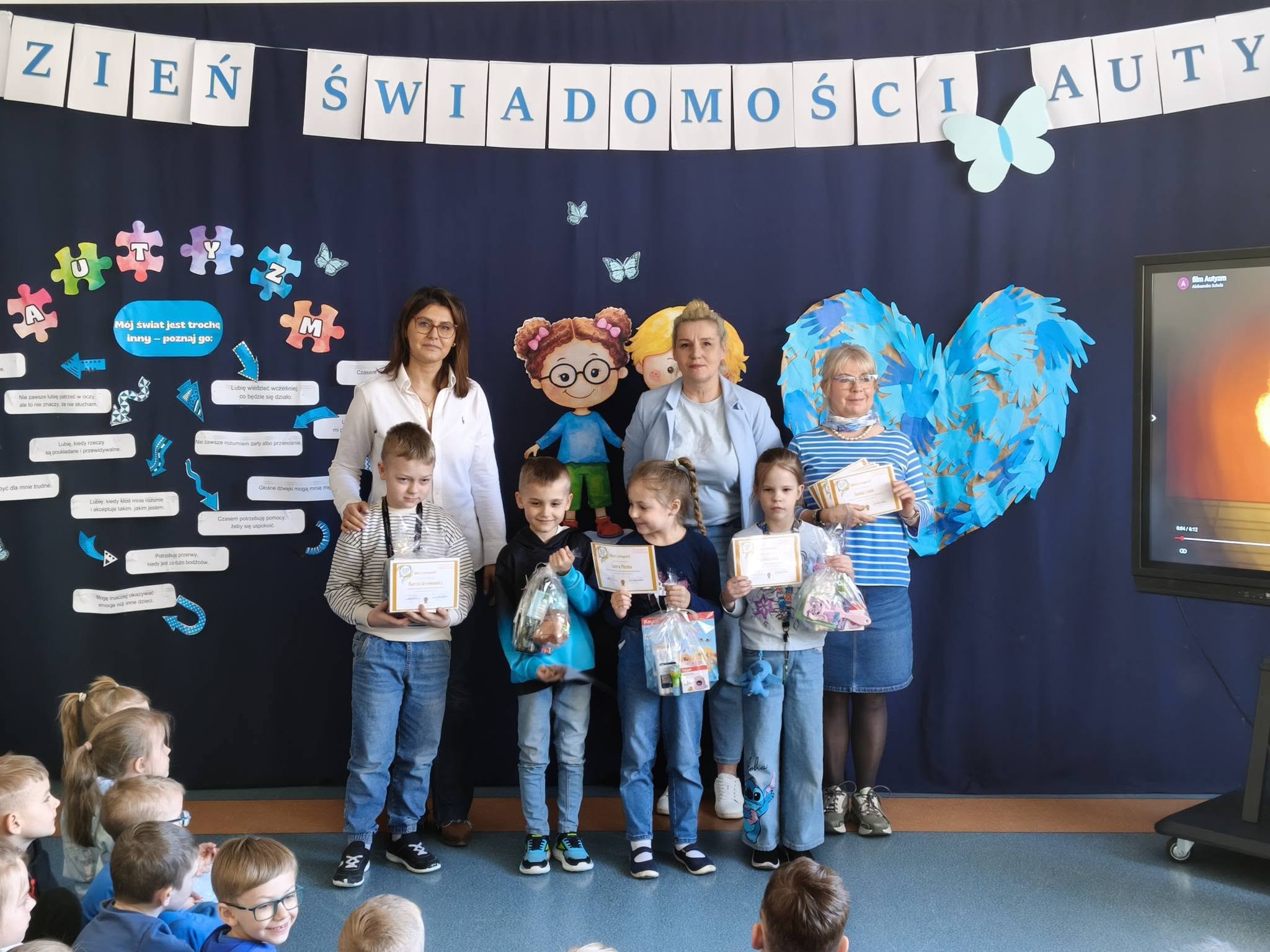 Dzieci z dyplomami i nagrodami na tle dekoracji z okazji Dnia Świadomości Autyzmu, z nauczycielkami.