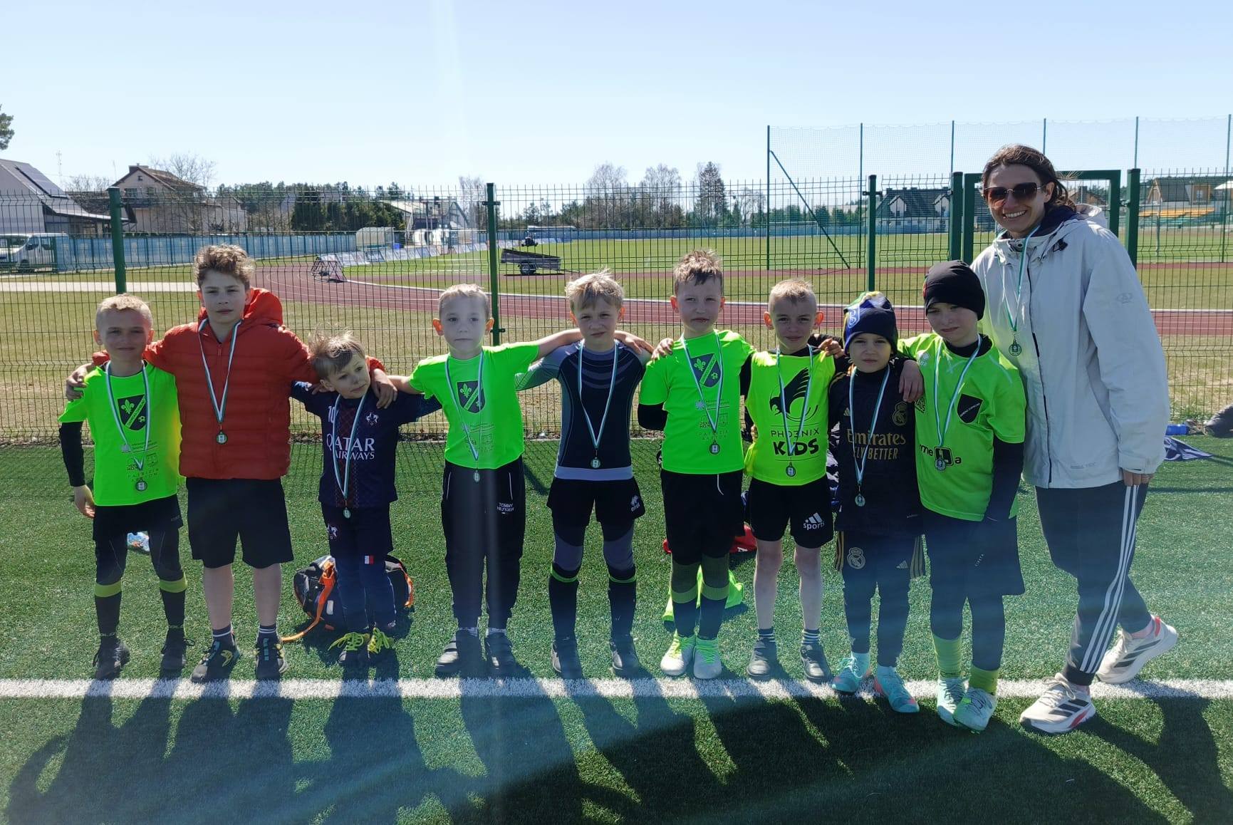 Drużyna dzieci z medalami i nauczycielką po zawodach sportowych na boisku.