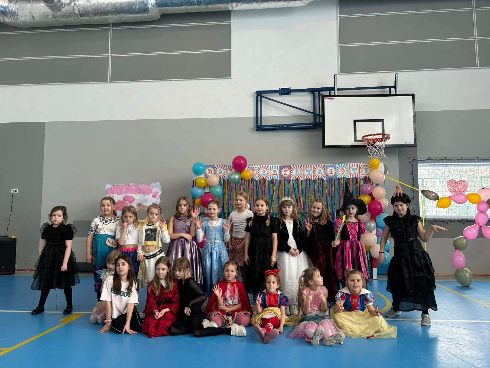 Grupa uczennic w kostiumach karnawałowych pozuje na tle dekoracji z balonów na sali gimnastycznej.
