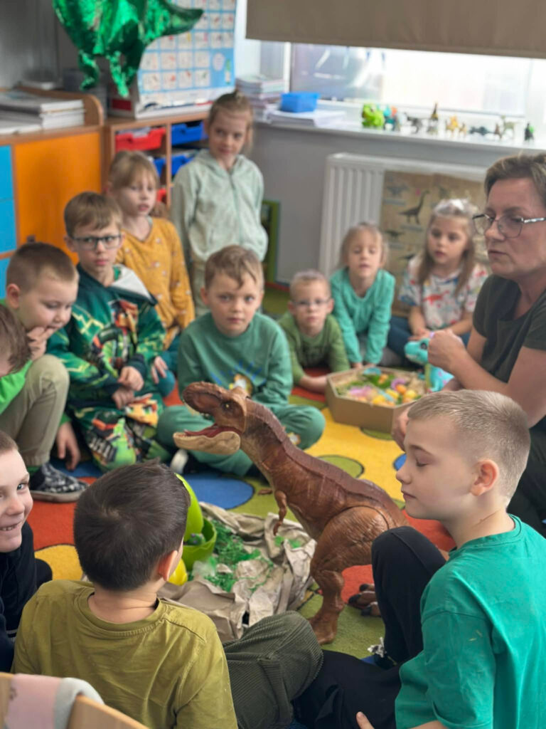 Grupa dzieci siedzi na kolorowym dywanie w kręgu, skupiona wokół dużej, realistycznej figurki Tyranozaura. Nauczycielka opowiada o życiu dinozaurów, a dzieci z zaciekawieniem obserwują makietę gniazda z jajem.