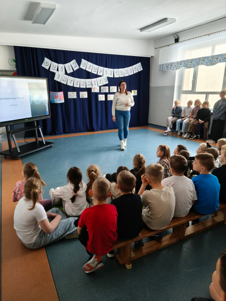 Uczniowie siedzący na ławeczkach słuchają prelekcji z okazji Dnia Bezpiecznego Internetu. W tle widoczna jest dekoracja tematyczna oraz ekran z prezentacją.
