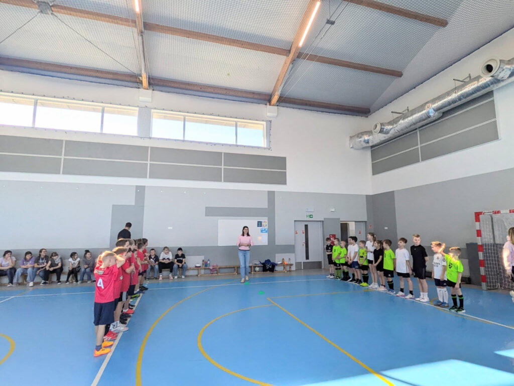 Uczniowie w strojach sportowych stoj膮 w dw贸ch rz臋dach na niebieskim parkiecie hali sportowej. Po艣rodku stoi nauczycielka prowadz膮ca zaj臋cia. Dzieci s膮 gotowe do rozpocz臋cia sportowych zmaga艅, a na 艂awkach z boku siedz膮 obserwuj膮cy wydarzenie koledzy.
