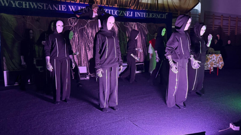Uczniowie ubrani w czarne bluzy z kapturem i białe maski wykonują układ taneczny na scenie. Nad nimi widnieje fragment napisu: „Zmartwychwstanie w epoce sztucznej inteligencji”. Scena jest oświetlona fioletowym, nastrojowym światłem.