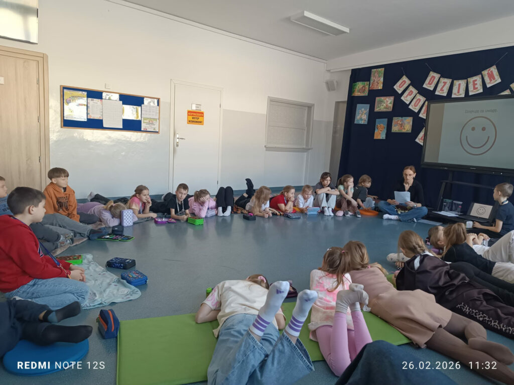Uczniowie siedzą i leżą w dużym kręgu na dywanie, słuchając nauczycielki, która czyta tekst. Na ekranie interaktywnym wyświetlona jest uśmiechnięta buźka, a sala udekorowana jest pracami plastycznymi.