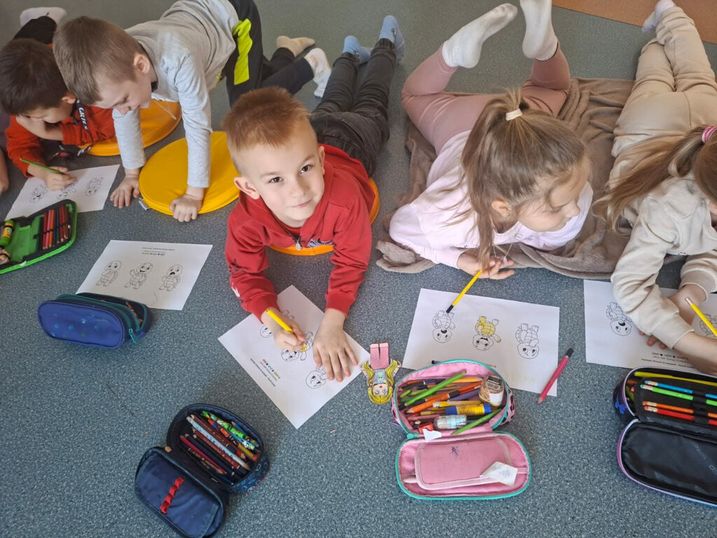 Uczniowie leżą na dywanie i kolorują karty pracy przedstawiające żółwie. Obok nich leżą otwarte piórniki z kredkami. Dzieci są skupione na zadaniu plastycznym.