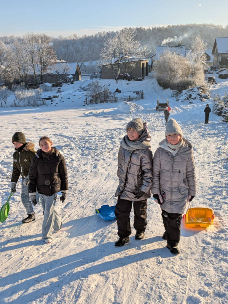 Czworo dzieci idzie pod górę ośnieżoną ścieżką, ciągnąc za sobą kolorowe sanki. W tle rozpościera się widok na wiejskie zabudowania i drzewa pokryte szronem pod bezchmurnym niebem.