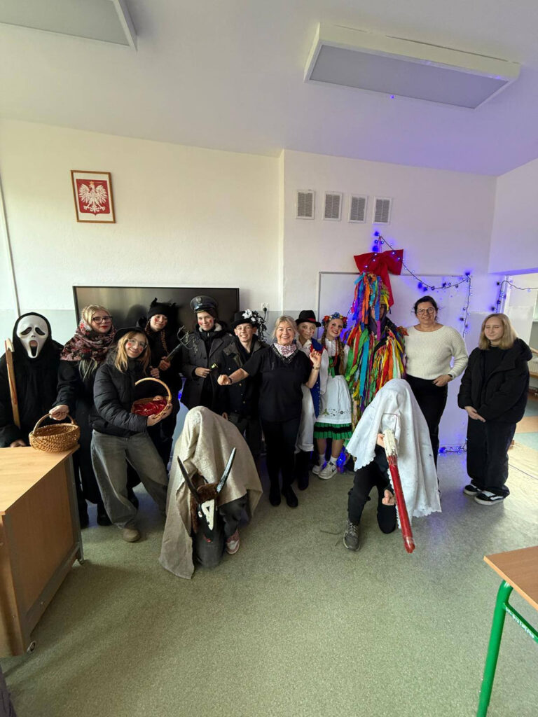 Kolędnicy w barwnych strojach pozują z nauczycielkami w udekorowanej sali lekcyjnej.