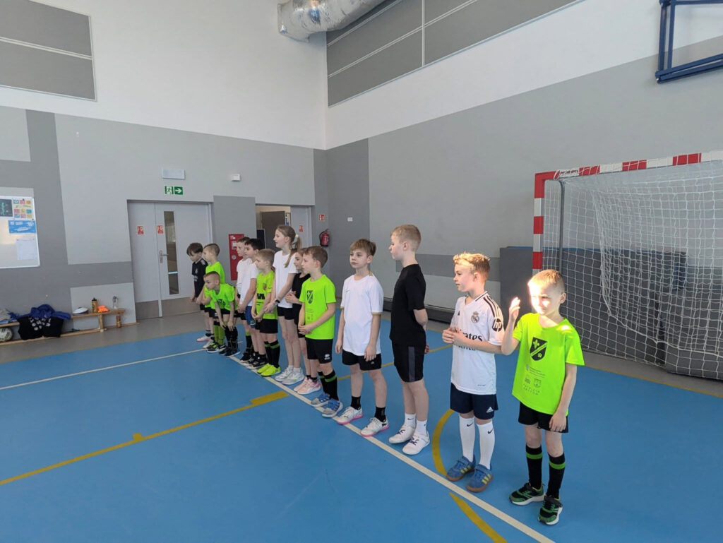 Grupa m艂odych sportowc贸w w neonowych, zielonych i bia艂ych koszulkach stoi w szeregu na boisku. Dzieci s膮 skupione i czekaj膮 na instrukcje trenera przed bramk膮 do pi艂ki r臋cznej