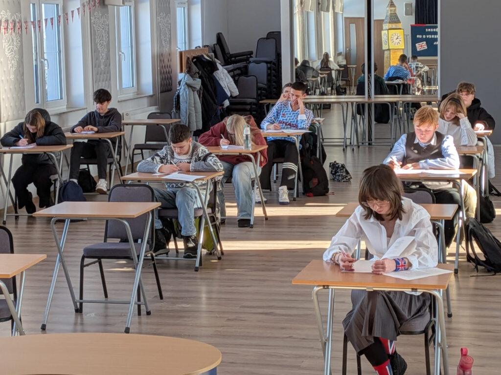 Uczniowie siedzą przy stolikach i rozwiązują test podczas finału konkursu wiedzy o Wielkiej Brytanii w szkolnej sali.