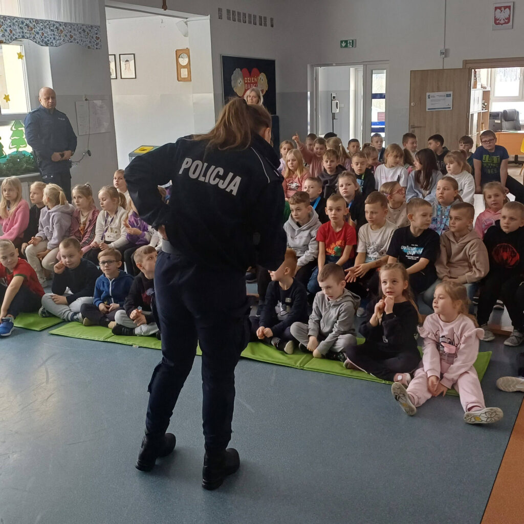Duża grupa uczniów siedzi na podłodze i materacach, słuchając funkcjonariuszki policji odwróconej tyłem do obiektywu. W tle widać drugiego policjanta oraz dekoracje szkolne.