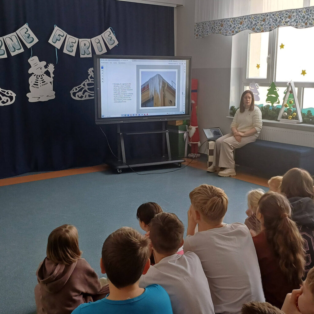 Dzieci siedzą tyłem do kamery, patrząc na ekran multimedialny, na którym wyświetlone jest zdjęcie dużych sopli lodu na dachu. Obok ekranu siedzi nauczycielka, a na oknie widać świąteczne ozdoby.