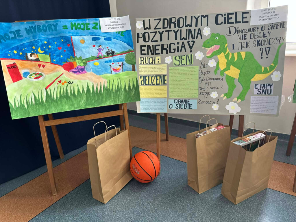 Wystawa nagrodzonych prac plastycznych wykonanych przez uczniów. Po lewej stronie barwny plakat o zdrowych wyborach, po prawej praca z dinozaurem i hasłem „W zdrowym ciele pozytywna energia”. Przed pracami stoją torby z nagrodami oraz piłka do koszykówki.
