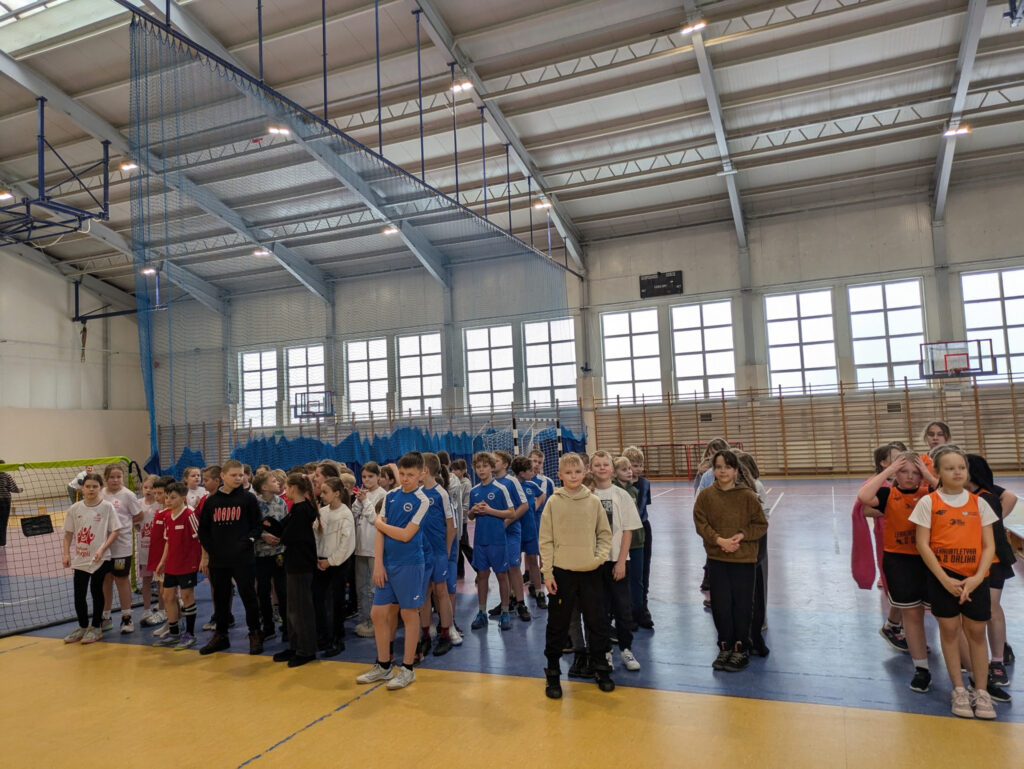 Grupy uczniów w strojach sportowych zebrane na hali podczas oficjalnego rozpoczęcia lub zakończenia zawodów.