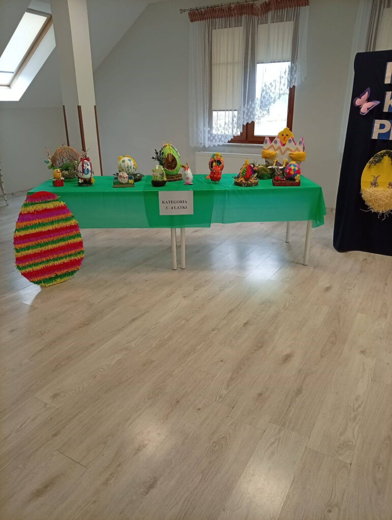 Stół z zielonym obrusem prezentujący prace plastyczne najmłodszych dzieci. Widoczne są m.in. kurczaki w skorupkach, ozdobne koszyczki i pisanki. Obok stołu na podłodze stoi duża, płaska dekoracja w kształcie wielobarwnej pisanki. Na stole tabliczka z napisem „KATEGORIA 3-4 LATKI”.