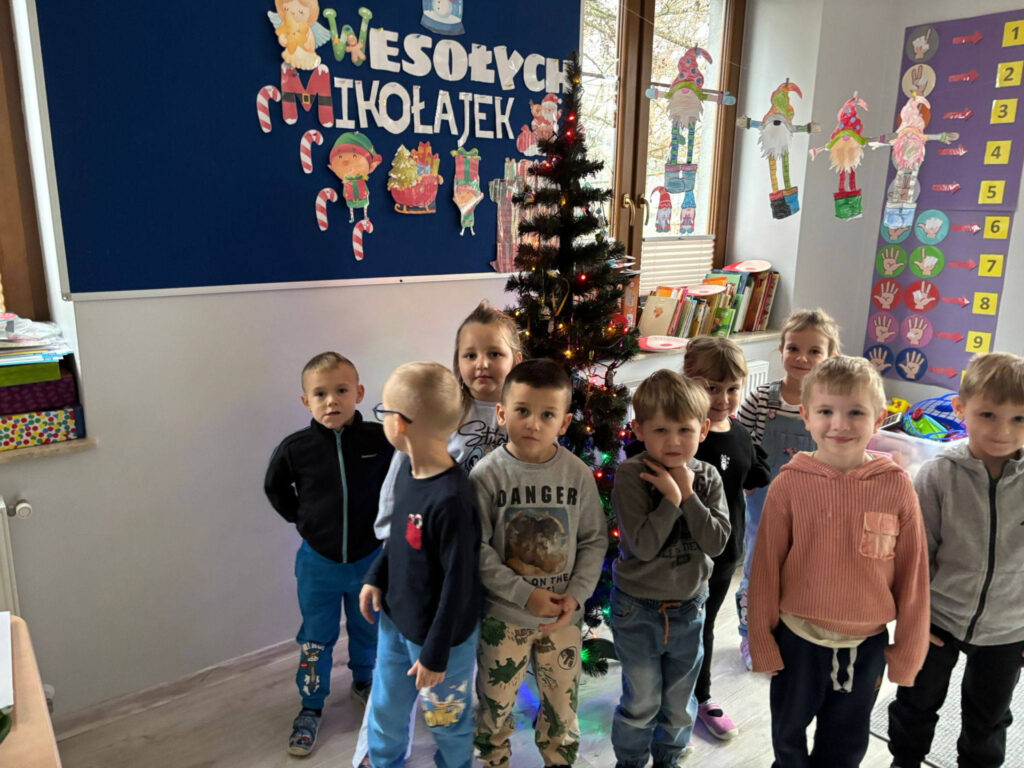 Grupa uczniów stoi w sali lekcyjnej przed choinką i niebieską gazetką ścienną z napisem „Wesołych Mikołajek”. Dzieci są ubrane w codzienne stroje, a obok nich widać okno oraz kolorowe pomoce dydaktyczne na ścianie.