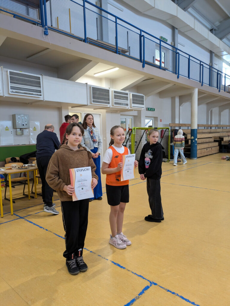 Dwie uczennice dumnie prezentują otrzymane dyplomy sportowe na podium sali gimnastycznej.