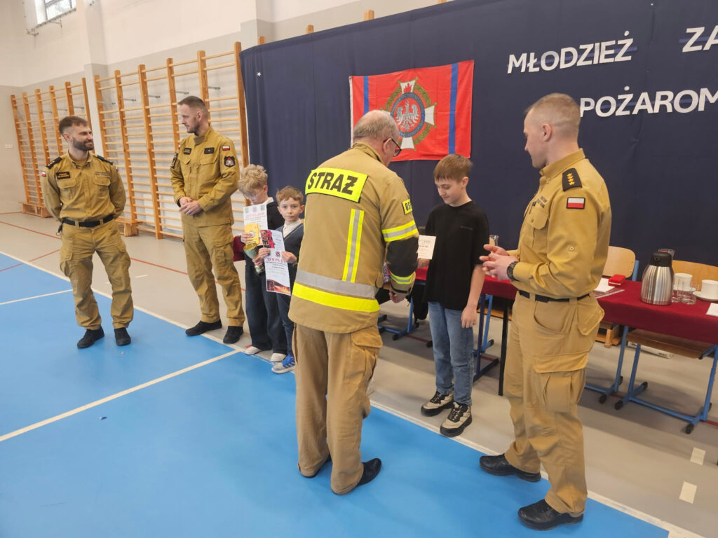 Scena wręczania nagród: jeden ze strażaków przekazuje dyplom uczestnikowi turnieju, podczas gdy pozostali strażacy i dzieci obserwują ten moment, stojąc obok stołu z nagrodami.
