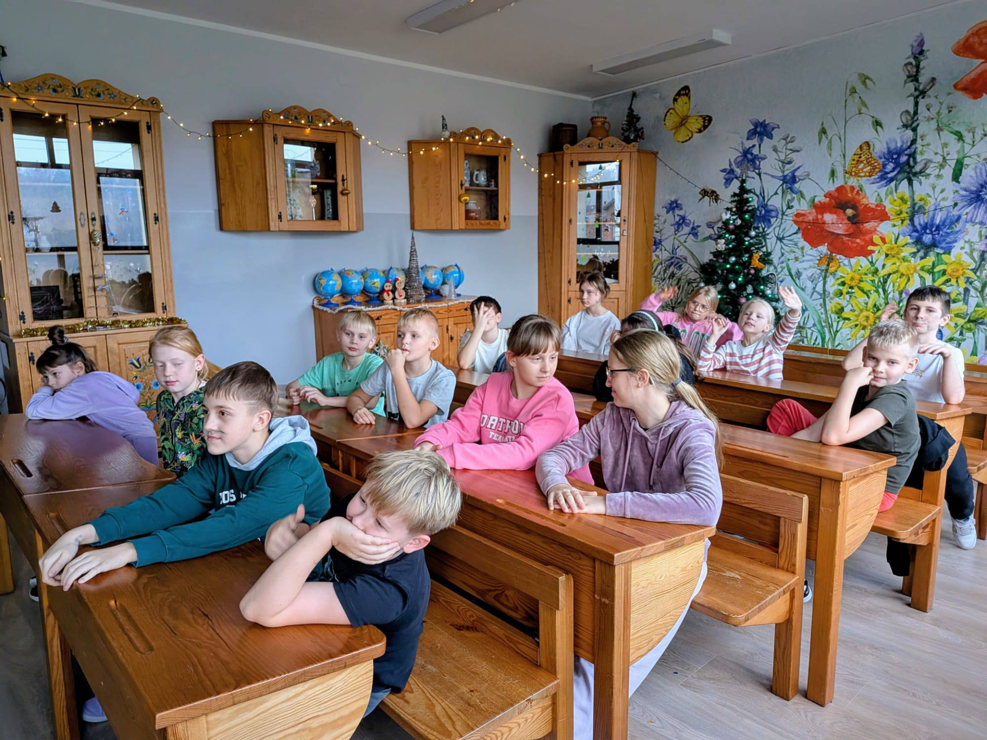 Grupa uczniów siedzi w drewnianych, klasycznych ławkach szkolnych w sali lekcyjnej.. Dzieci patrzą w stronę kamery lub rozmawiają ze sobą. W tle widać ścianę z kolorowym muralem w kwiaty, choinkę oraz szafki ozdobione światełkami.