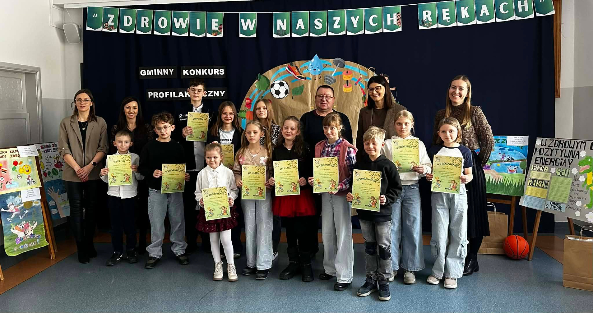 Laureaci Gminnego Konkursu Profilaktycznego „Zdrowie w naszych rękach” pozują do wspólnego zdjęcia z nauczycielami. Uczniowie z dumą prezentują otrzymane dyplomy i wyróżnienia. W tle widać dekorację z hasłem konkursu oraz rysunkami promującymi sport i zdrowe żywienie.