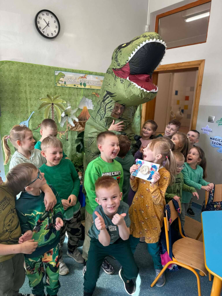Radosna grupa dzieci pozuje do zdjęcia z osobą przebraną w wielki, dmuchany kostium dinozaura. Uczniowie śmieją się, niektórzy pokazują kciuki w górę, a jedna z dziewczynek prezentuje swoją pracę plastyczną. W tle widać dekorację z napisem „Dzień Dinozaura”.