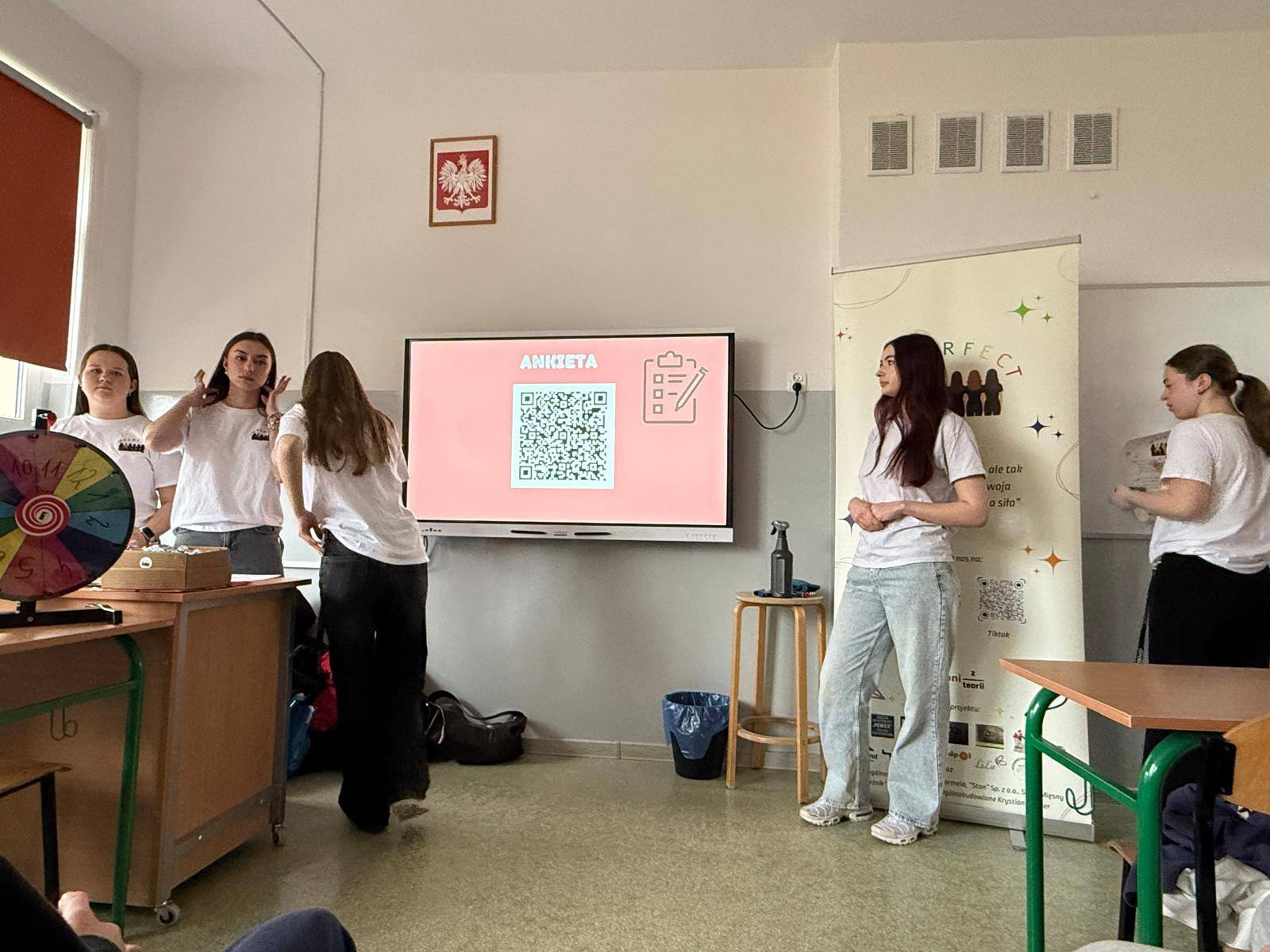 Uczennice w białych koszulkach prezentują projekt „Ankieta” na ekranie multimedialnym.