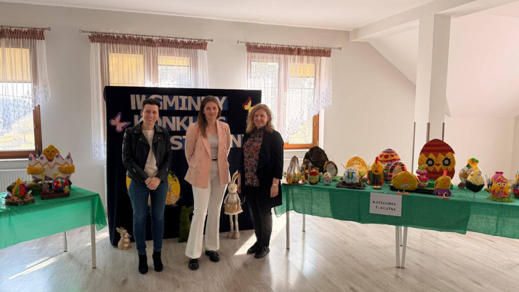 Trzy uśmiechnięte kobiety (prawdopodobnie komisja konkursowa lub nauczycielki) pozują na tle wystawy prac wielkanocnych. Stoją przed czarną tablicą z napisem „IV GMINNY KONKURS...”. Po obu stronach widoczne są stoły z kolorowymi dekoracjami wykonanymi przez dzieci.
