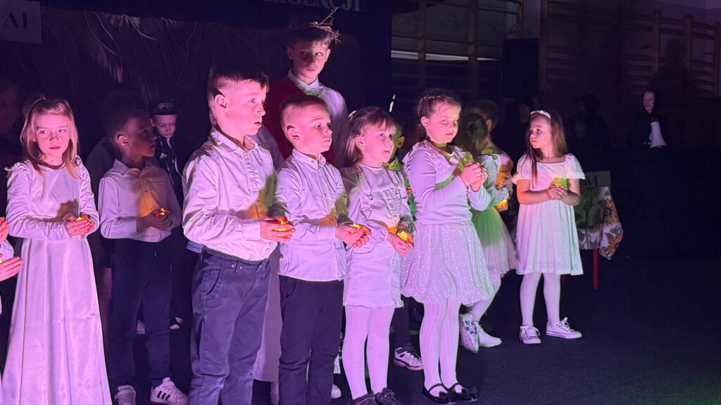 Grupa dzieci w jasnych, galowych strojach stoi na zaciemnionej scenie podczas występu. Każde dziecko trzyma w dłoniach małe, palące się światełko.