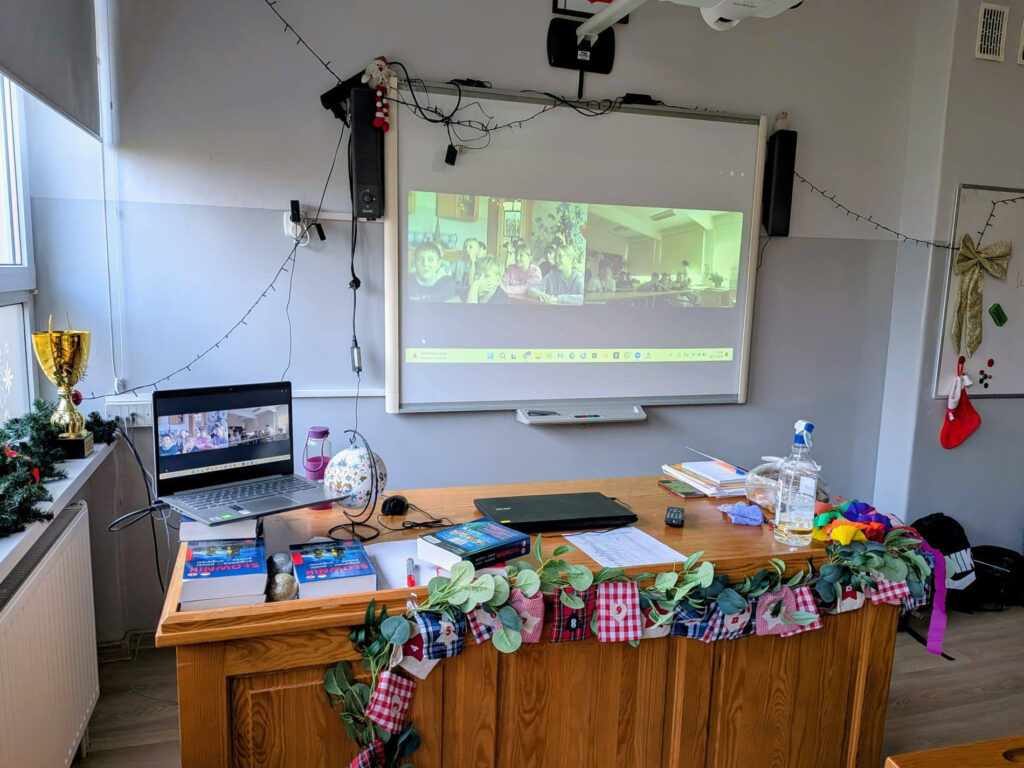 Klasa przygotowana do wideokonferencji. Na drewnianym biurku, udekorowanym świątecznymi girlandami i kalendarzem adwentowym, stoi otwarty laptop i słowniki polsko-angielskie. W tle widać białą tablicę z rzutnikiem wyświetlającym obraz z połączenia wideo z inną grupą dzieci.