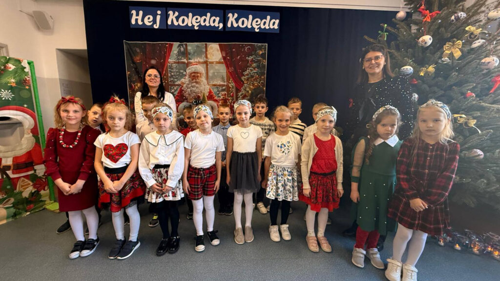 Dzieci w strojach galowych i ludowych akcentach stoją pod napisem „Hej Kolęda, Kolęda” obok choinki.
