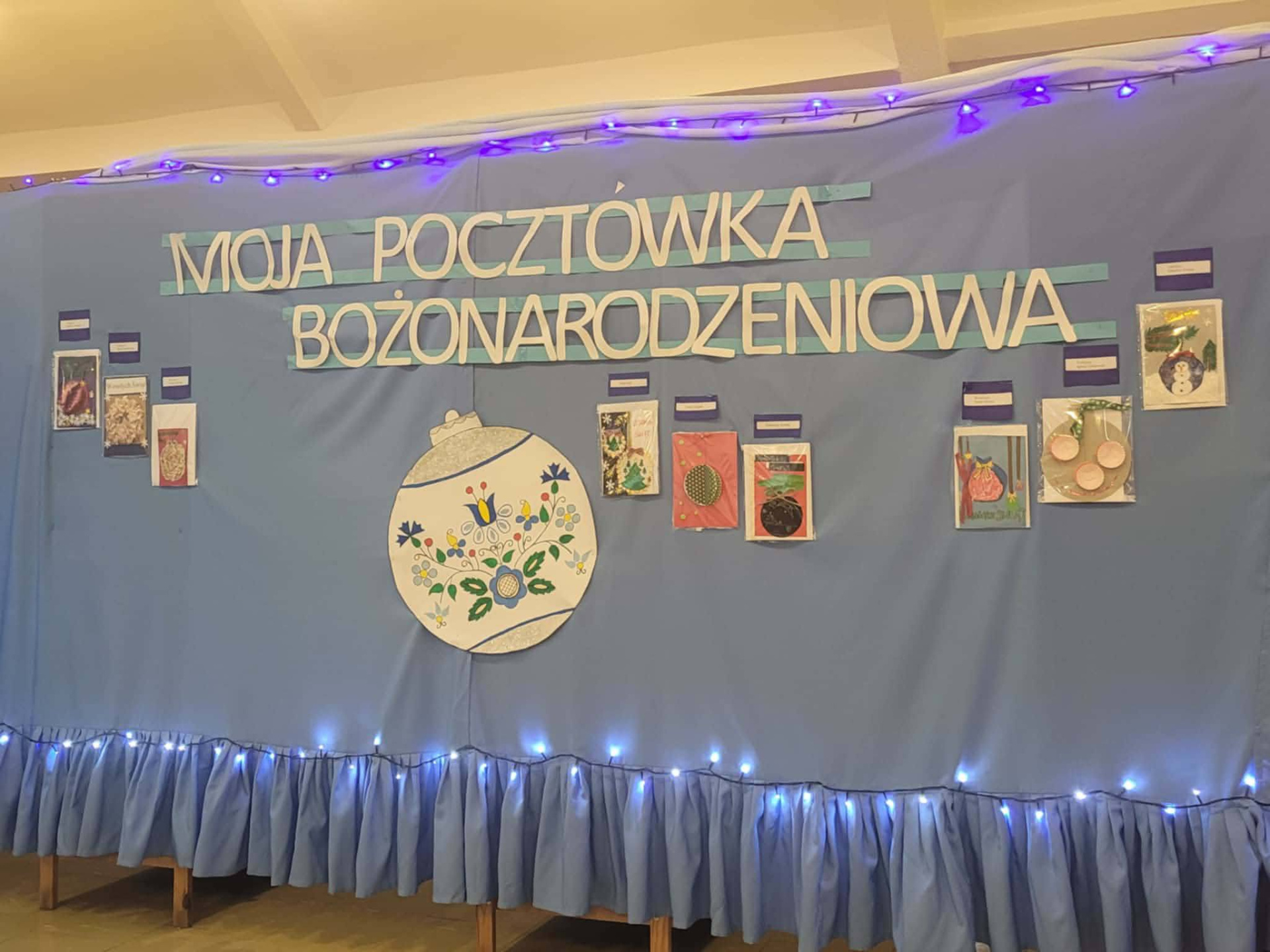 Niebieska ścianka z napisem „Moja Pocztówka Bożonarodzeniowa”, ozdobiona pracami plastycznymi dzieci i lampkami.