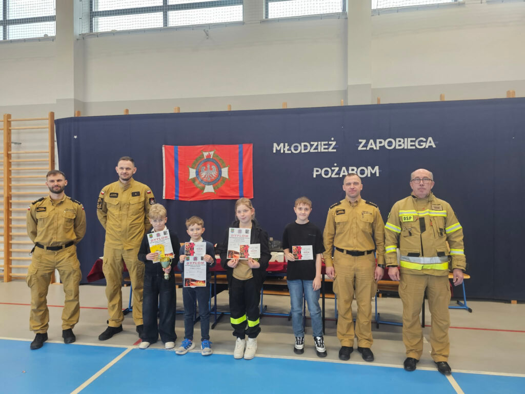 Oficjalne zdjęcie laureatów turnieju pożarniczego: czworo dzieci prezentujących dyplomy i puchary w otoczeniu czterech strażaków w mundurach służbowych na tle flagi i banneru.