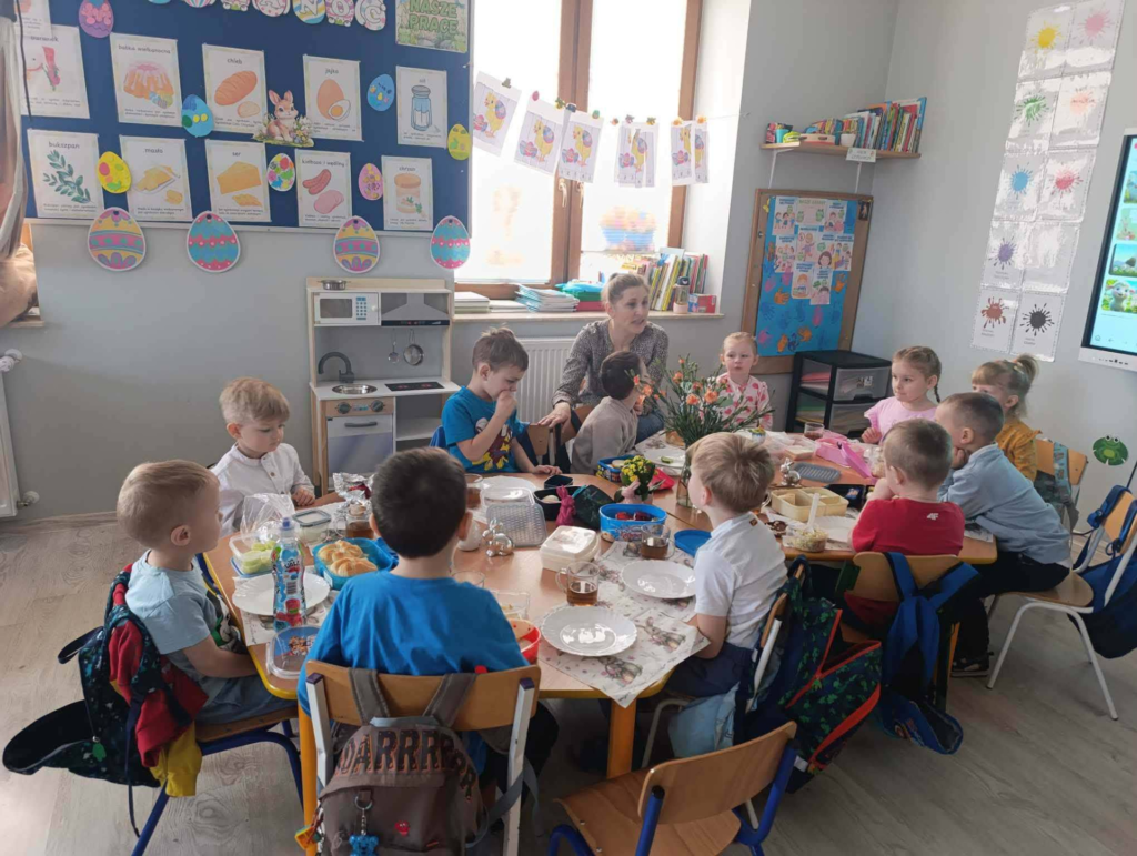 Grupa dzieci w wieku przedszkolnym siedzi wraz z nauczycielk膮 przy du偶ym wsp贸lnym stole nakrytym do 艣niadania. Dzieci rozmawiaj膮 i jedz膮 posi艂ek w jasnej sali lekcyjnej.