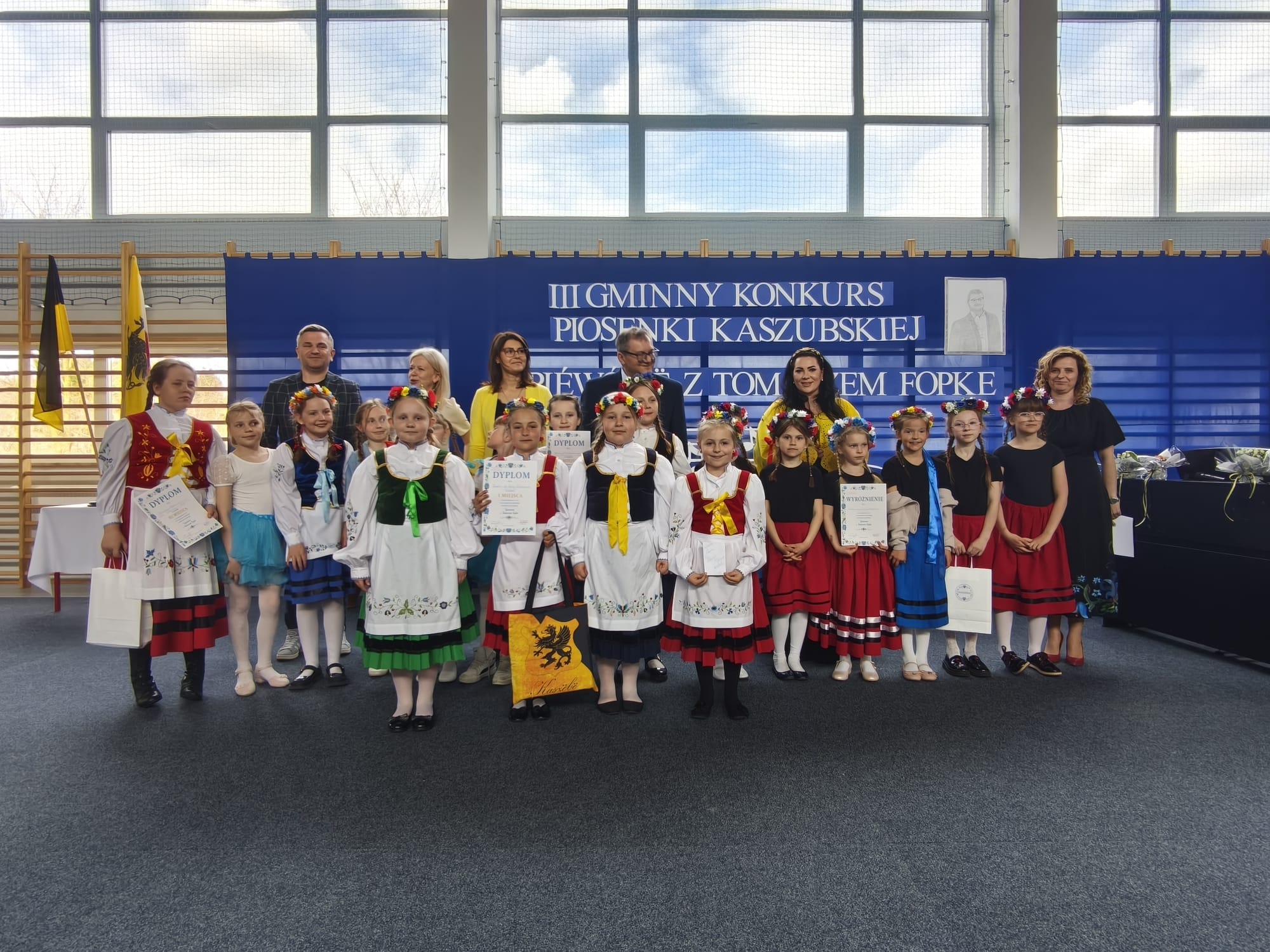 Najmłodsze dzieci w kolorowych strojach regionalnych pozują do zdjęcia z dyplomami i opiekunami na tle sceny konkursowej.