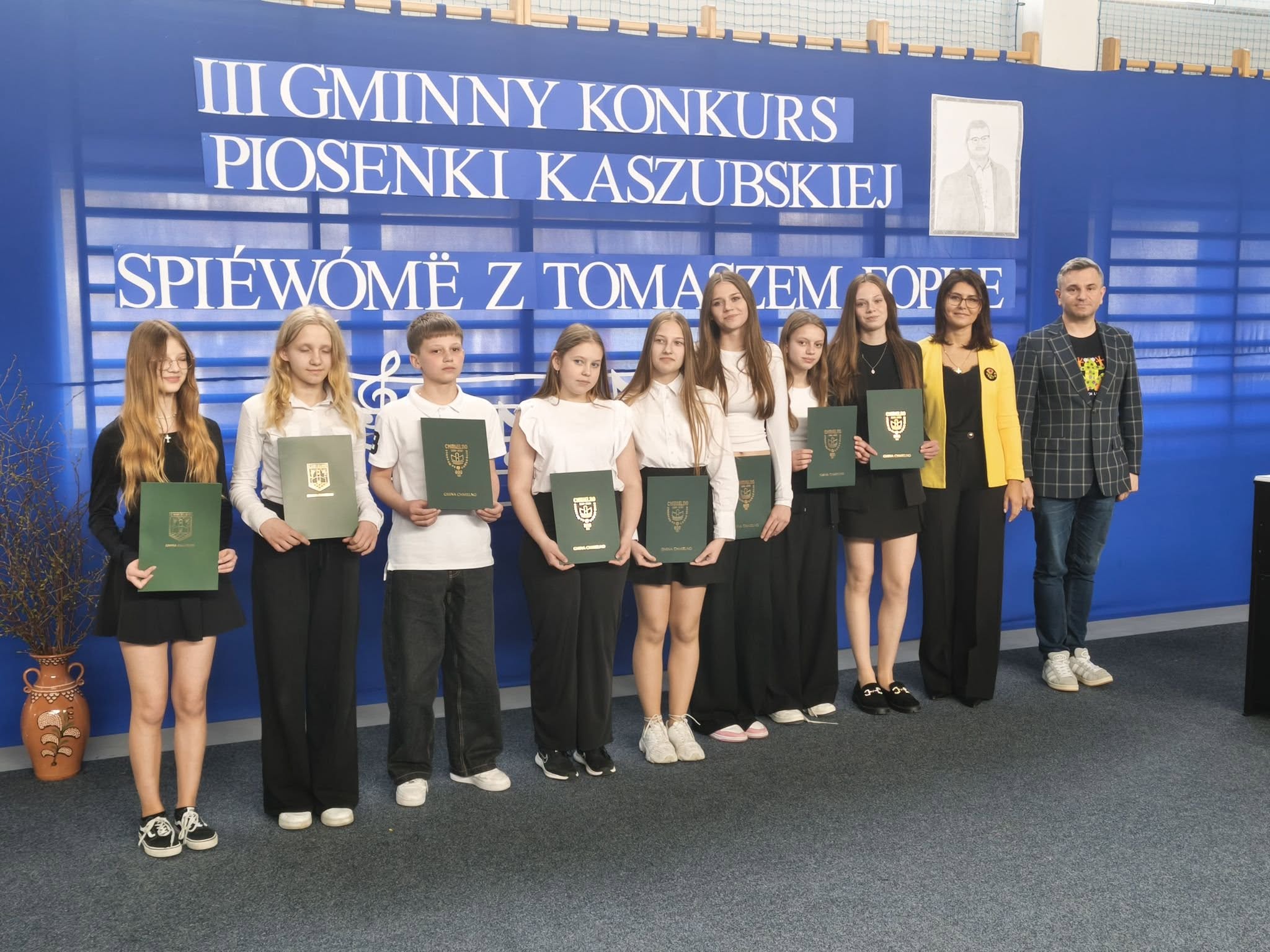 Grupa uczniów stoi na tle sceny konkursowej, trzymając stypendia i pozując do zdjęcia z dyrektorem szkoły- Martą Grzenia oraz Wójtem Gminy Chmielno- Panem Michałem Melibrudom