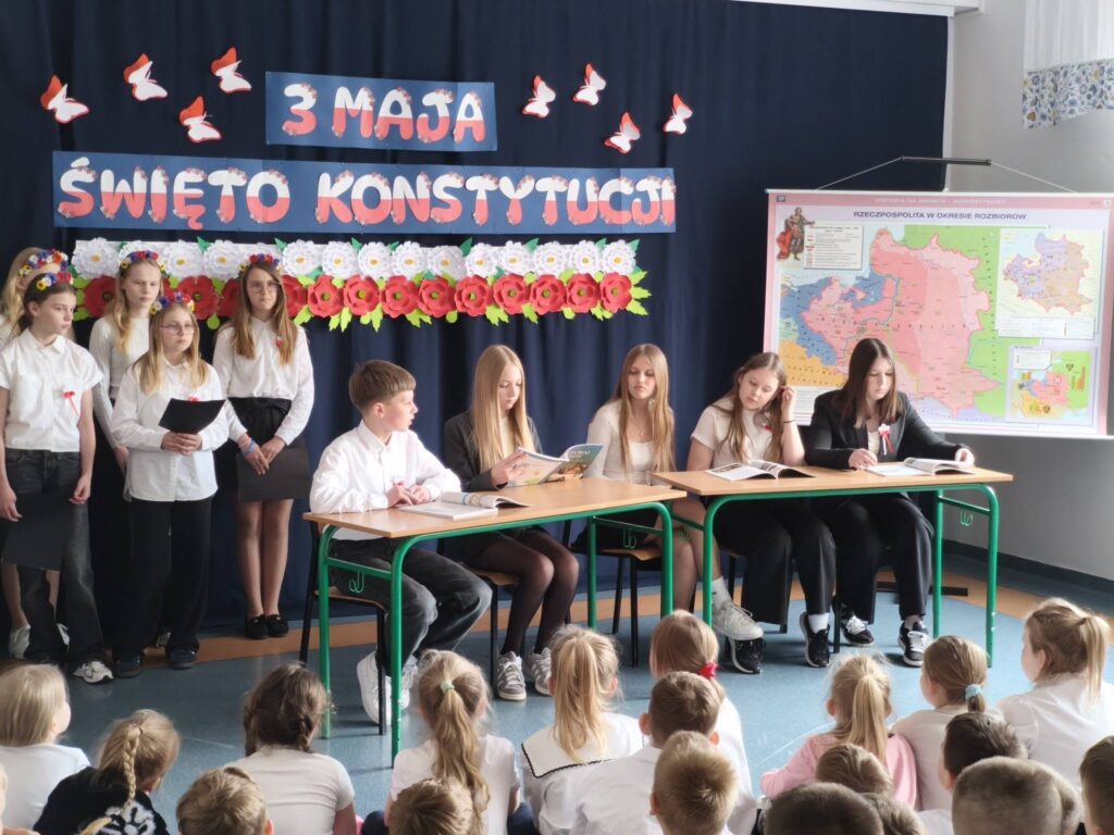 Uczniowie siedzą przy ławkach i stoją obok, prezentując program artystyczny z okazji Święta Konstytucji 3 Maja przed młodszą publicznością.