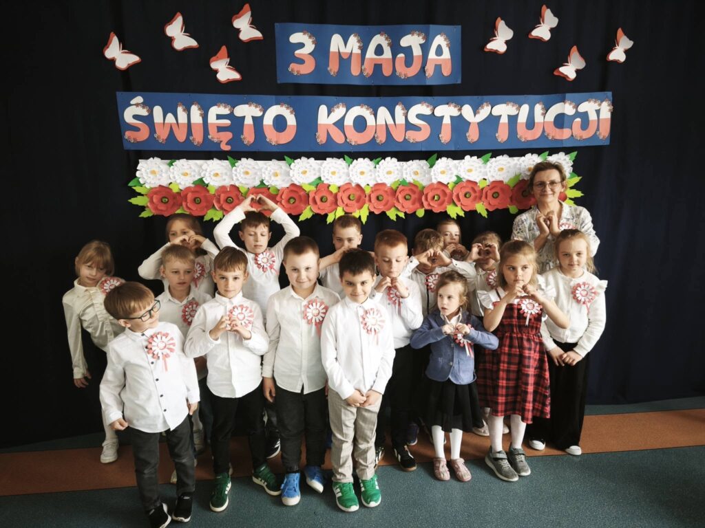 Najmłodsze dzieci wraz z nauczycielką pozują do zdjęcia na tle dekoracji „3 Maja – Święto Konstytucji”, wykonując gest serca rękami.