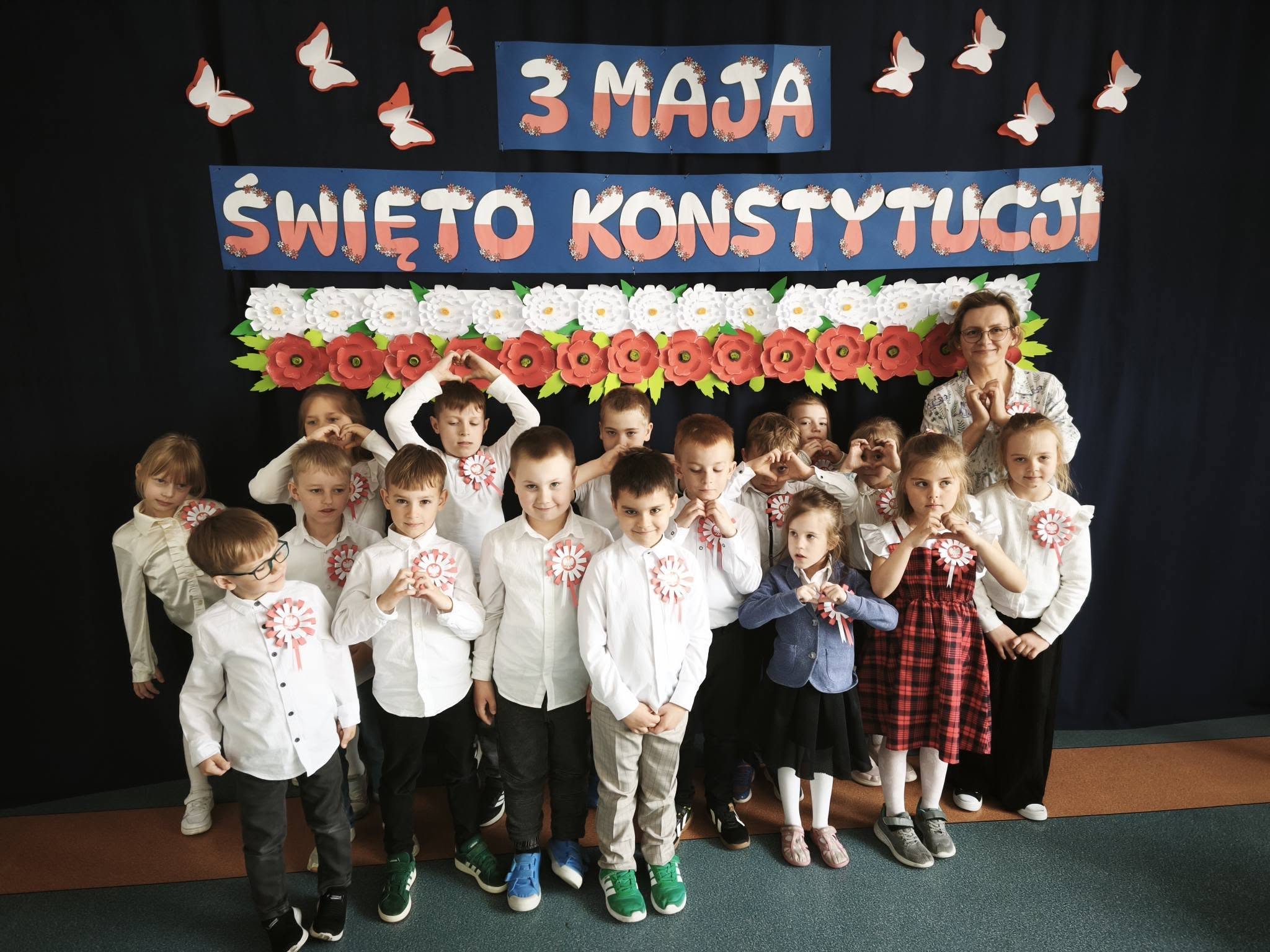 Najmłodsze dzieci wraz z nauczycielką pozują do zdjęcia na tle dekoracji „3 Maja – Święto Konstytucji”, wykonując gest serca rękami.