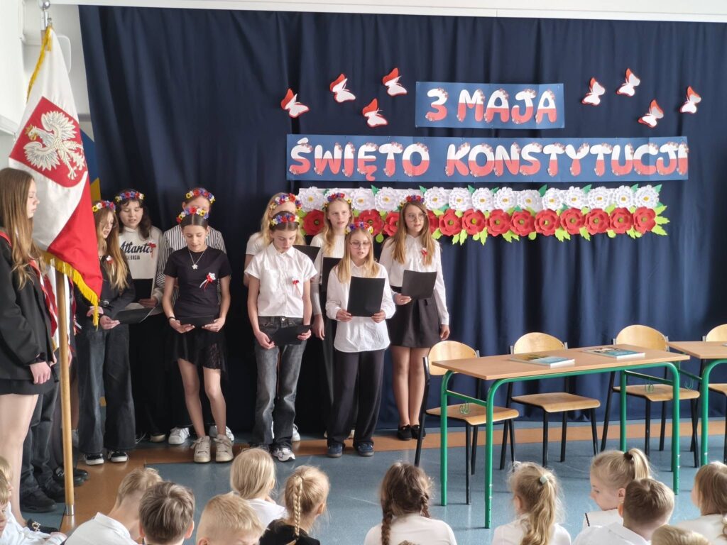 Grupa uczniów stoi na scenie z kartkami w rękach, śpiewając podczas szkolnej uroczystości z okazji 3 Maja.