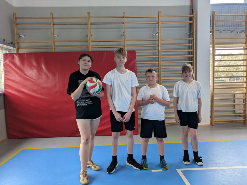 Czworo uczni贸w pozuje w sali gimnastycznej podczas szkolnych zaj臋膰 sportowych. Jedna osoba trzyma pi艂k臋 do siatk贸wki, a w tle widoczne s膮 materace i drabinki gimnastyczne.