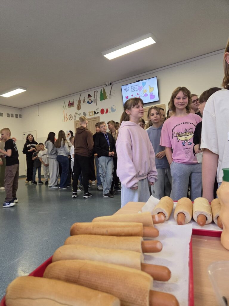 Uczniowie stoj膮 w szkolnym korytarzu podczas kiermaszu lub szkolnego wydarzenia. Na pierwszym planie wida膰 hot dogi przygotowane na tacy, a w tle grupy dzieci rozmawiaj膮 i ustawiaj膮 si臋 przy stoiskach.