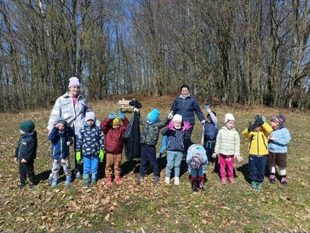 Grupa przedszkolaków wraz z opiekunkami stoi na świeżym powietrzu podczas wspólnej akcji sprzątania terenu. Dzieci ubrane są w kolorowe kurtki i czapki, a jedna z osób trzyma worek na zebrane śmieci.