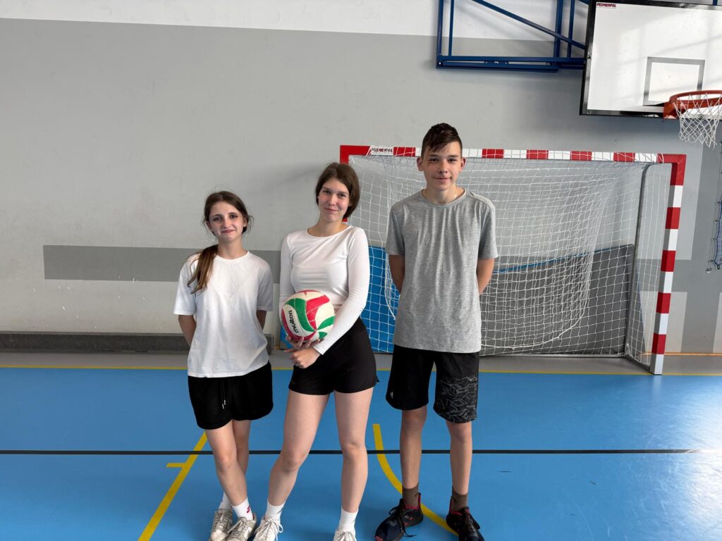 Trzyosobowa dru偶yna uczni贸w stoi w szkolnej sali gimnastycznej przed bramk膮 do pi艂ki r臋cznej. Jedna z uczennic trzyma pi艂k臋 do siatk贸wki, a wszyscy maj膮 sportowe stroje.