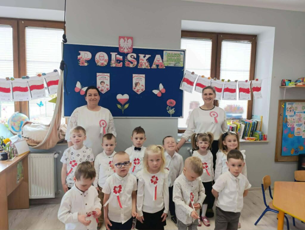 Grupa przedszkolaków i nauczycielek pozuje w sali udekorowanej symbolami narodowymi z okazji święta Polski. Dzieci ubrane są na biało i mają przypięte biało-czerwone kotyliony.