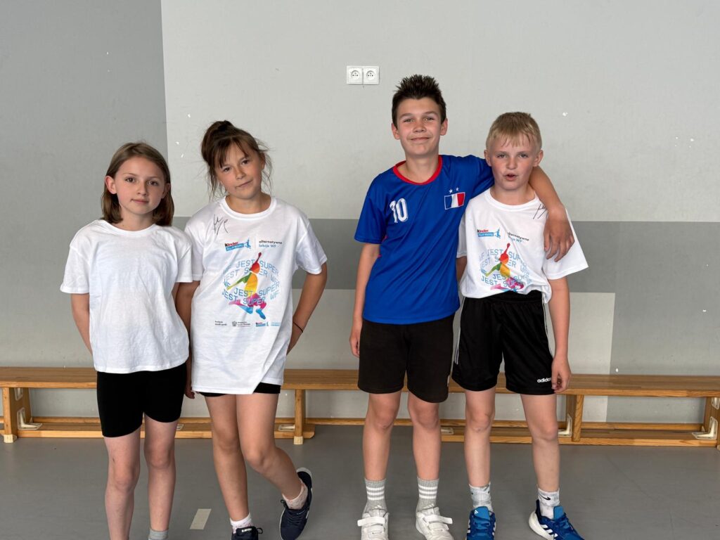 Grupa czworga dzieci stoi razem w sali gimnastycznej po szkolnych zawodach lub zaj臋ciach sportowych. Uczniowie ubrani s膮 w stroje sportowe i pozuj膮 do wsp贸lnego zdj臋cia.