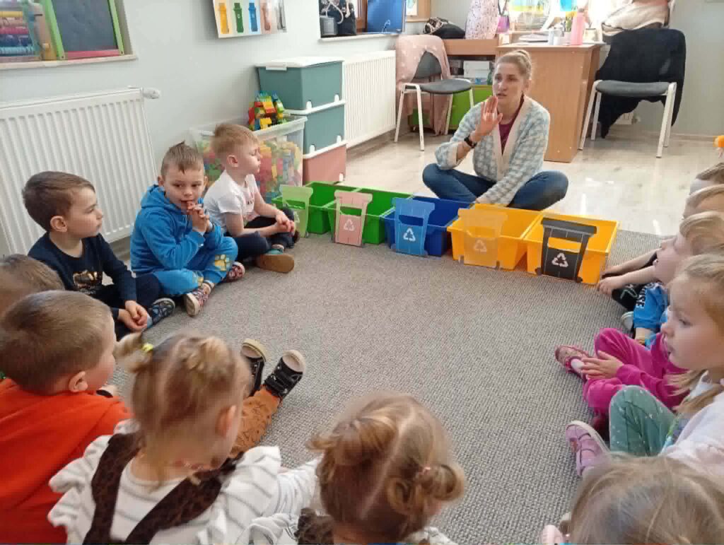 Dzieci siedzą w kręgu w sali przedszkolnej podczas zajęć edukacyjnych o segregacji odpadów. Nauczycielka pokazuje kolorowe pojemniki do recyklingu i rozmawia z dziećmi.