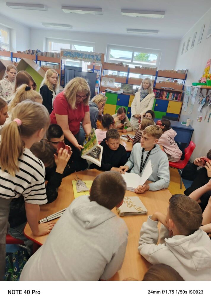 Grupa uczniów i nauczycieli zebrana wokół stołu podczas zajęć w szkolnej bibliotece.