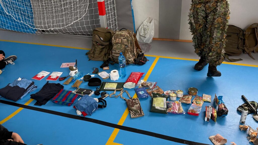 Na podłodze sali gimnastycznej rozłożony jest sprzęt survivalowy i wyposażenie potrzebne podczas wypraw terenowych. Widoczne są plecaki, żywność, odzież, apteczka i różne akcesoria biwakowe.