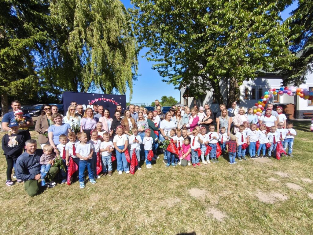 Dzieci, rodzice i nauczyciele podczas rodzinnej uroczystości plenerowej pozują do wspólnego zdjęcia na terenie szkoły.