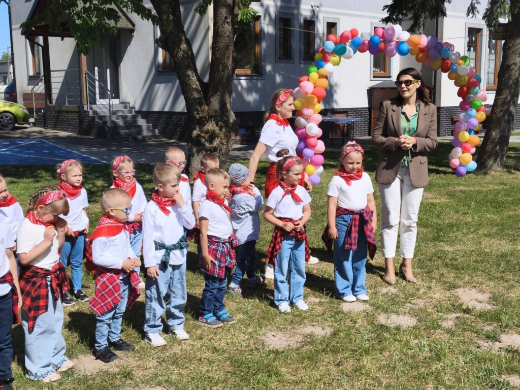 Przedszkolaki występują podczas uroczystości rodzinnej, a nauczycielka prowadzi wydarzenie na szkolnym dziedzińcu.