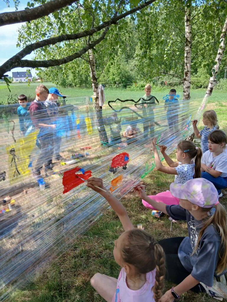 Dzieci tworzą wspólną pracę plastyczną, malując na rozciągniętej folii podczas zajęć integracyjnych na świeżym powietrzu.